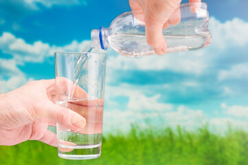 Hands holding plastic bottle and glass with pure water on grassland landscape background