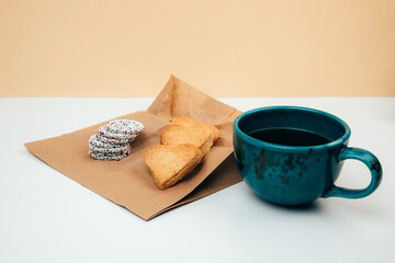 A cup of coffee with foam and a plate with heart-shaped shortbread cookies and chocolate rounds on a paper bag. Front view