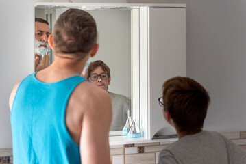 Dad and son in bathroom in front of mirror together. Young father and his teenage son with shaving foam on his nose