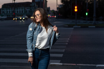 Obraz premium Brunette girl in youth clothes on evening city background. Stylish young woman in glasses on city street, summer time