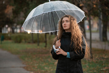 Sadness attractive woman with long hair in autumn park in rainy weather under transparent umbrella. Portrait of beautiful girl