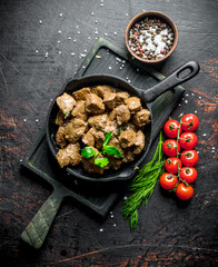 Pieces of cooked liver in a frying pan on a cutting Board with tomatoes, herbs and spices.