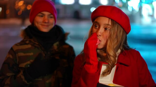 Teen Caucasian girl posing eating delicious sweets as blurred boy smiling standing at background. Positive teenage couple dating in night city on Valentine's looking at camera
