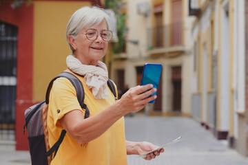 Fototapeta premium Carefree senior traveler woman carrying backpack visiting the old town of Seville looking at phone, smiling elderly lady enjoying travel and discovery