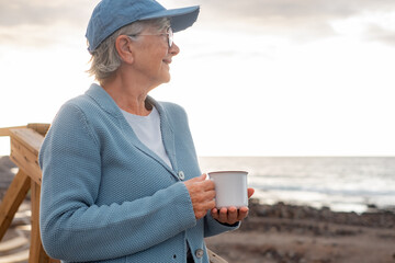 Relaxed handsome senior woman with cap and eyeglasses standing outdoor at sunrise holding a cup of coffee or tea. Smiling elderly lady enjoying silence of the morning on the beach