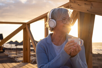 Relaxed handsome senior woman with headphones listening music while sitting outdoor at sunrise...