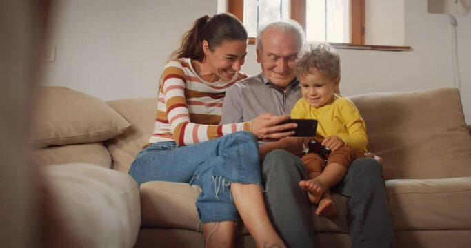 Happy Small Family Gathered Together, Sitting on a Sofa in the Living Room and Using a Smartphone. Mother, Son and Grandfather Watching Videos on a Mobile Phone and Laughing. They are Bonding 