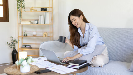 Beautiful Asian woman working on laptop and sipping coffee with smiling face in her home, Creating happiness at work with a smile, Freelancer working at home happily, Work from home.