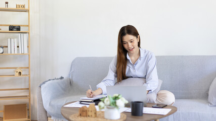 Obraz na płótnie Canvas Beautiful young asian woman watching live video or video call of teacher teaching on laptop in her home, Take notes of important conversations and messages during the teacher's teaching.