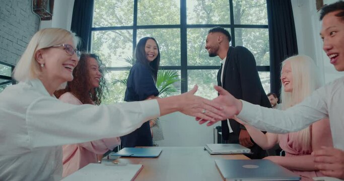 Happy multiethnic business people celebrate partnership agreement shaking hands at office table meeting slow motion.