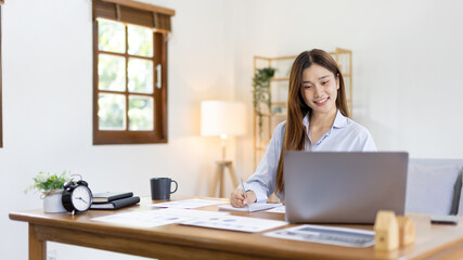 Beautiful young asian woman watching live video or video call of teacher teaching on laptop in her home, Take notes of important conversations and messages during the teacher's teaching.