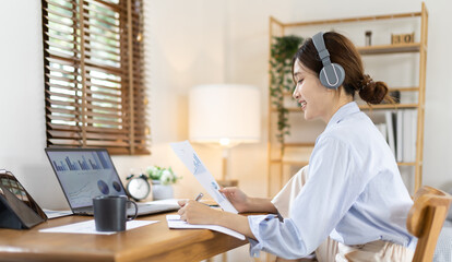 Naklejka na ściany i meble Asian female college student wearing headphones watching live performance or video call teacher teaching on laptop, Conversations with teachers and classmates, Online learning, Study at your own home.