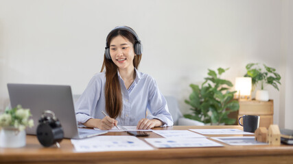 Asian female college student wearing headphones watching live performance or video call teacher teaching on laptop, Conversations with teachers and classmates, Online learning, Study at your own home.