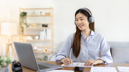 Asian female college student wearing headphones watching live performance or video call teacher teaching on laptop, Conversations with teachers and classmates, Online learning, Study at your own home.