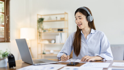 Asian female college student wearing headphones watching live performance or video call teacher teaching on laptop, Conversations with teachers and classmates, Online learning, Study at your own home.