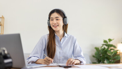 Asian female college student wearing headphones watching live performance or video call teacher teaching on laptop, Conversations with teachers and classmates, Online learning, Study at your own home.