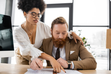 seductive african american businesswoman hugging bearded coworker and pointing at document on desk.