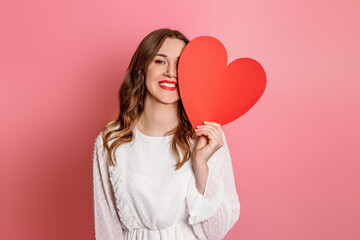 girl closes one eye with a big paper heart isolated on a pink background in the studio. Valentine's...