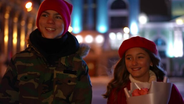 Dolly Shot Portrait Of Romantic Teenage Couple Talking Strolling Outdoors And Leaving. Front View Happy Loving Caucasian Teen Boyfriend And Girlfriend Dating In Night City On Valentine's Walking Away