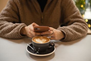 Girl in a fluffy sweater keeps smartphone in hands and writes message next to cup of coffee with heart shaped latte art foam. Closeup cup of coffee with cream in coffee shop. St. Valentine's Day. Love