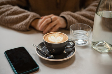 Girl in cafe waits for call from loved one over cup of coffee with heart shaped latte art foam. Closeup cup of coffee with cream in coffee shop. St. Valentine's Day. Love
