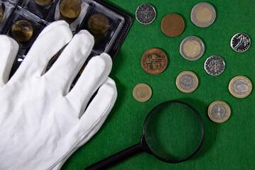 Coins on a green cloth, white gloves and a magnifying glass, top view.