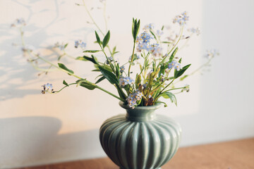 Beautiful little blue flowers in vase in warm sunlight on rustic wooden background. Delicate myosotis petals, forget me not spring flowers. Atmospheric evening moment. Simple countryside living
