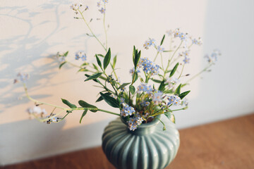 Beautiful little blue flowers in vase in warm evening sunlight on rustic wooden table. Delicate myosotis petals, forget me not spring flowers. Simple countryside living, home decor