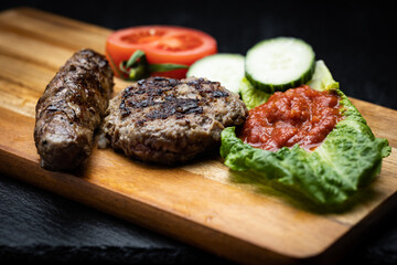 Traditional Bulgarian homemade grilled ground meat sausages called kyufte and kebapche placed on wooden plank next to dipping sauce lyutenica and slices of cucumber and tomato
