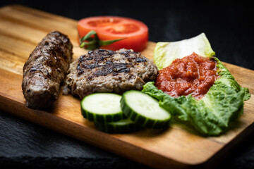Traditional Bulgarian grilled ground meat meals kyufte and kebapche garnished with cucumber and tomato slices next to lyutenica dipping sauce