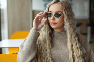 Naklejka premium Fashion beautiful blonde girl model in a beige sweater wears vintage stylish sunglasses and sits in a cafe