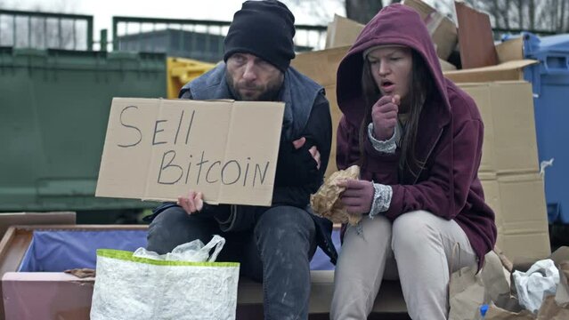 Two dirty and poorly dressed homeless people, a man and a woman, sit by a pile of rubbish with a handwritten SELL BITCOIN poster. Young woman is sick and coughing.