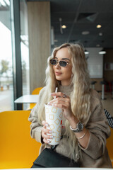 Trendy beautiful fashion blond hipster woman with sunglasses in a beige sweater holds a paper cup of cola and sits in a cafe