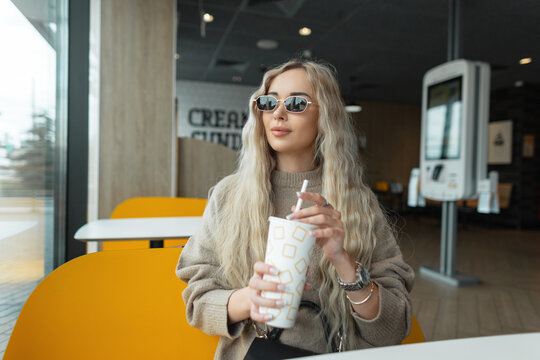 Happy beautiful blond hipster girl with vintage sunglasses in a fashion beige sweater sits in a cafe and drinks cola from a paper cup