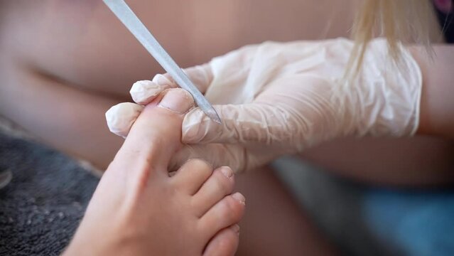 Podiatrist master in white gloves using sterile hardware file to polish toe nail and create suitable shape, therapeutic pedicure in beauty salon, beautician cleaning patient's feet and nails before
