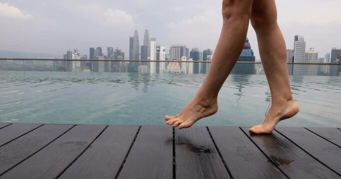 Bare female legs walk along wooden side of open-air pool on roof of skyscraper. Woman steps on tiptoes along dark decking next to pool. Relax by pool on roof of tall skyscraper in heart of city