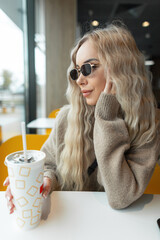 Beautiful young hipster woman with blond curly hairstyle with vintage sunglasses in a fashion beige sweater sits in a cafe and holds a paper cup of cola