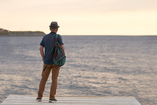 An Older, Gray-haired Man With A Hat And A Backpack Behind His Back Looks Out From The Pier. Copy Space. Selective Focus.