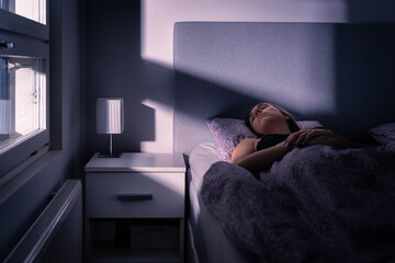 Sleeping woman in bed at night in dark bedroom. Person under blanket and cover. Person asleep, resting in cold room at home. Tired lady in peaceful dream. Blue moonlight from window. Insomnia.