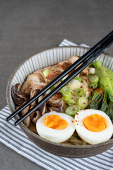 Asian noodle soup, ramen with chicken breast, vegetables, mushrooms and egg in the bowl on a striped napkin and gray concrete background. Front view from above, healthy eating concept
