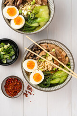 Two bowls of ramen soup with chicken breast, vegetables, mushrooms, pak choi and egg on a white wooden table and chopsticks. Top view, healthy eating concept