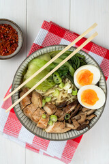 Bowl of ramen soup with chicken breast, vegetables, mushrooms and egg on a red checked napkin on a white wooden table and chopsticks. Top view, healthy eating concept