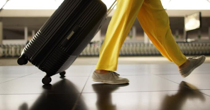A woman in transit zone of airport rushes to next flight to make a transfer and travel to another country. A close view of woman's legs in bright yellow pants. A woman is dragging suitcase on wheels.