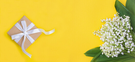 Gift box with tied white bow and bouquet of lily of the valley on the yellow background. Top view. Copy space.