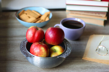 Stack of books, e-reader, reading glasses, bowl of biscuits, apples and cup of tea on the table. Bookshelf in the background. Selective focus.