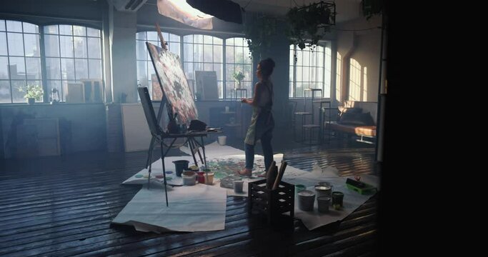 Woman artist picks up paint from can and walks over to canvas to continue working on abstract painting on large canvas in spacious studio. Female artist creates modern masterpiece with hands and oil.