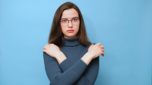Displeased brunette student girl in glasses and a sweater embraces herself, holds shoulders, feels cold, missed opportunity, expresses regret, dreams of warm and cozy home on blue background