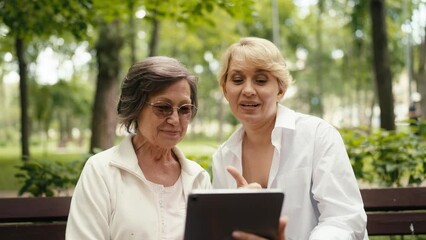 Happy elderly woman and her friend using tablet while sitting on a park bench