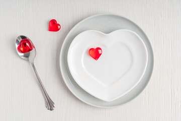 Table top view of empty ceramic heart-shaped plate decorated with bright red heart decor served on white wooden background with silver spoon. Tableware prepared for celebration of valentines day