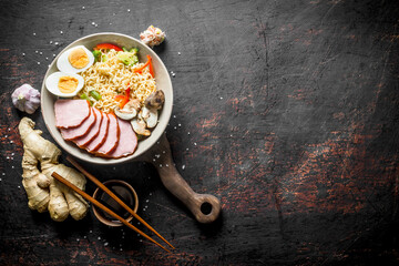 Instant noodles in bowl with ham, vegetables and egg on cutting Board.